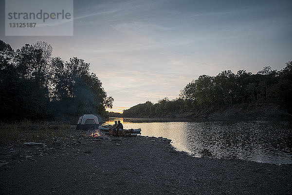 Männliche Freunde sitzen bei Sonnenuntergang auf einem Baumstamm auf einem Campingplatz am See gegen den Himmel