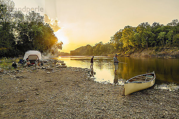 Junger Mann sitzt auf dem Campingplatz  während Freunde bei Sonnenuntergang im See angeln