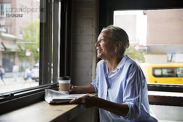 Lächelnde Frau mit Zeitung und Kaffee  die durch das Fenster am Tisch im Café schaut