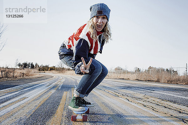 Porträt einer verspielten Frau  die ein Friedenszeichen gestikuliert  während sie im Winter mit dem Skateboard auf der Strasse unterwegs ist