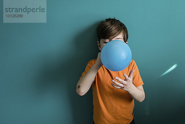 Junge bläst Ballon auf  während er zu Hause an blauer Wand steht