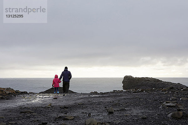 Rückansicht eines Vaters mit Tochter auf einem Felsen bei Dyrholaey Island vor bewölktem Himmel