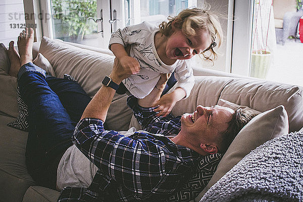 Glücklicher Vater hebt Tochter hoch  während er zu Hause auf dem Sofa liegt