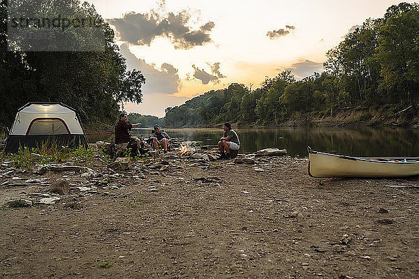 Männliche Freunde unterhalten sich  während sie bei Sonnenuntergang am Campingplatz gegen den Himmel sitzen