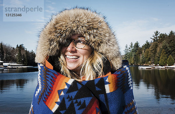 Fröhliche Frau trägt Pelzmantel  während sie in eine Decke gewickelt am See im Algonquin-Provinzpark liegt