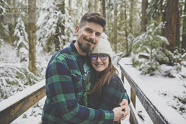 Porträt eines lächelnden Paares  das sich im Winter im Lynn Canyon Park auf einem Steg inmitten von Bäumen im Wald umarmt