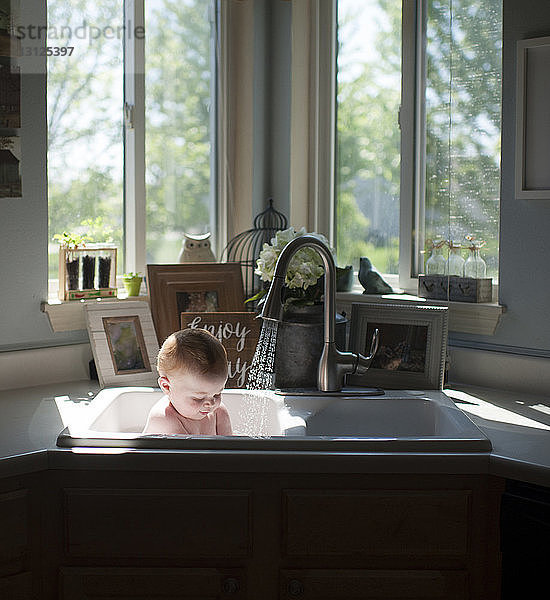 Kleiner Junge ohne Hemd  der zu Hause in der Spüle vor dem Fenster sitzt