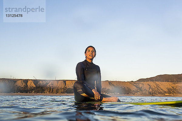 Junge Frau sitzt auf einem Surfbrett im Meer vor klarem Himmel
