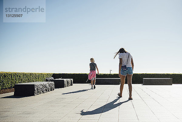 Rückansicht von Mädchen  die bei strahlendem Sonnenschein auf einem Fußweg gegen den klaren Himmel laufen