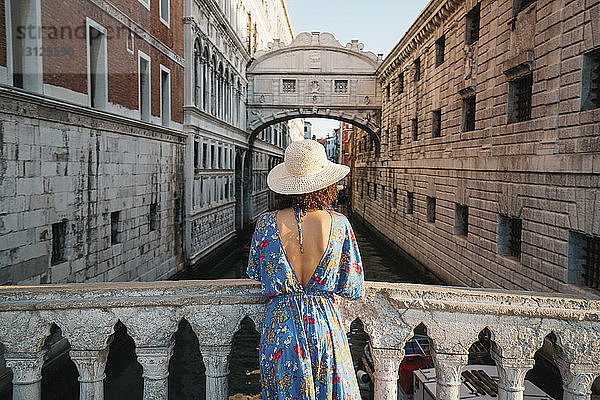 Rückansicht einer Frau mit Hut  die an einem Geländer auf einer Brücke inmitten von Gebäuden in der Stadt steht