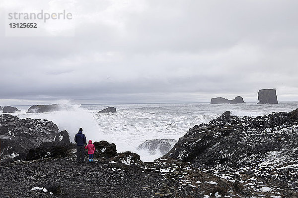Rückansicht eines Vaters mit Tochter  der auf das Meer schaut  während er auf einem Felsen bei Dyrholaey Island vor bewölktem Himmel steht