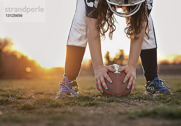 Niedriger Teil eines Mädchens  das American Football in der Hand hält  während es bei Sonnenuntergang auf einem Rasenfeld gegen den Himmel steht