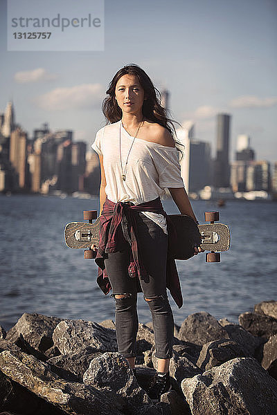 Porträt einer Frau  die ein Skateboard auf einer felsigen Küste gegen den Himmel hält