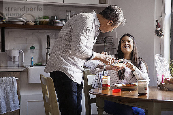 Lächelndes Mädchen sieht Vater an  der Erdnussbutter auf Brot in der Küche aufträgt