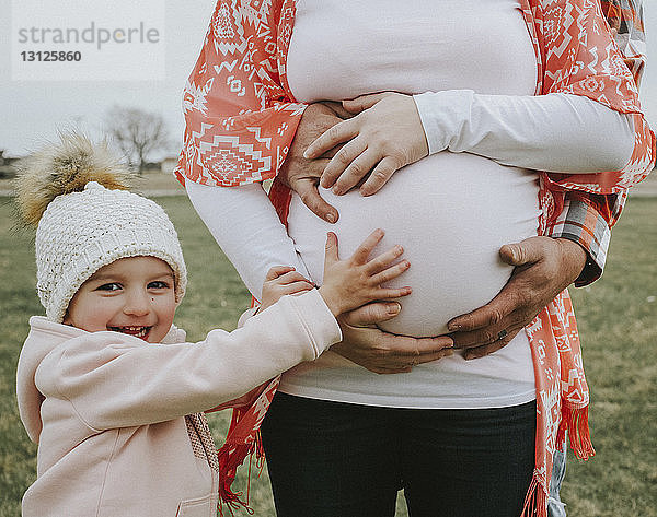 Porträt einer fröhlichen Tochter  die den Bauch der schwangeren Mutter berührt  mit dem Vater auf dem Feld