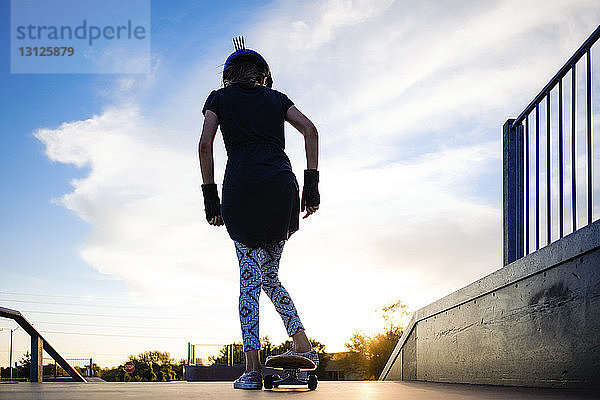 Rückansicht eines Skateboard fahrenden Mädchens vor bewölktem Himmel im Park bei Sonnenuntergang