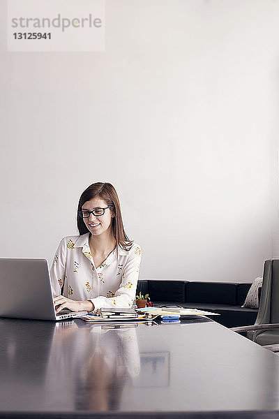 Selbstbewusste Geschäftsfrau benutzt Laptop  während sie am Tisch an der Wand sitzt