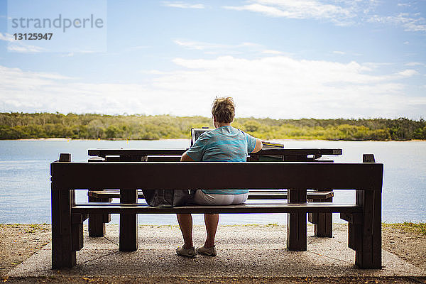 Rückansicht einer älteren Frau mit einem Laptop  die auf einer Bank am Seeufer sitzt und gegen den Himmel gerichtet ist