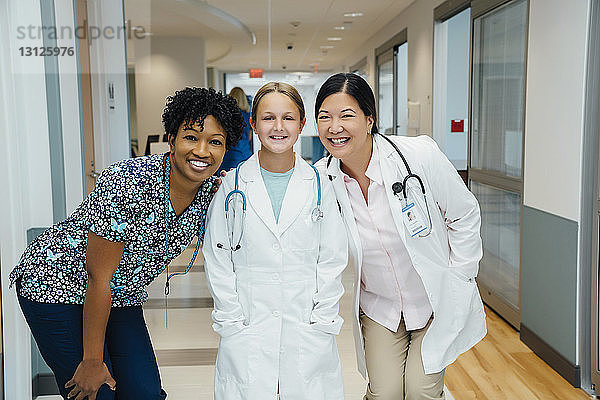 Porträt von fröhlichen Ärztinnen mit Mädchen im Laborkittel im Krankenhauskorridor