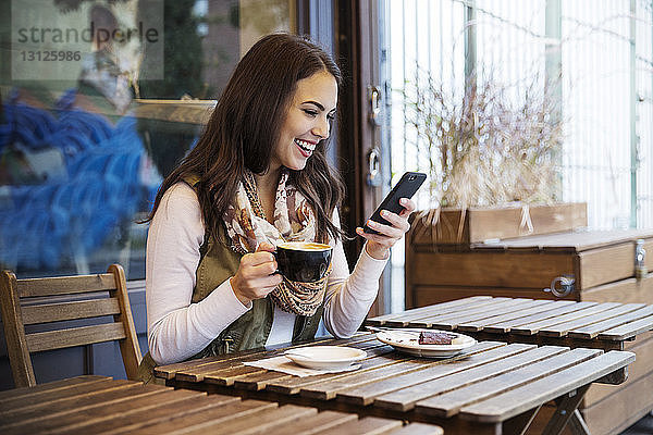 Lächelnde Frau hält Kaffeetasse in der Hand  während sie im Café ein Mobiltelefon benutzt