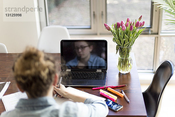 Rückansicht einer Frau mit Laptop-Computer  die zu Hause auf einem Buch schreibt