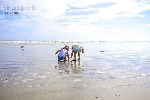 Geschwister spielen im nassen Sand am Strand gegen den Himmel