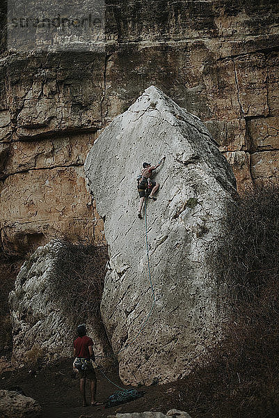 Rückansicht eines Mannes  der ein Seil hält  während ein Freund im Wald am Fels klettert