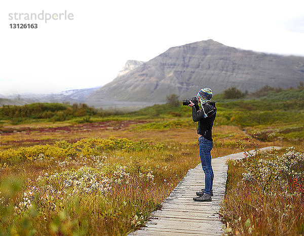 Seitenansicht eines Wanderers  der mit der Kamera fotografiert  während er auf einer Strandpromenade vor Bergen steht