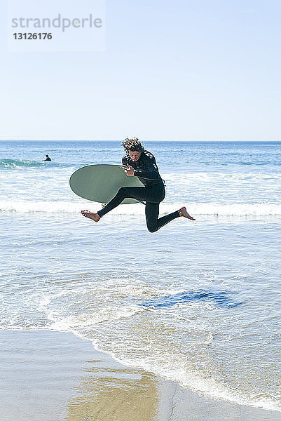 Mann mit Surfbrett springt im Meer gegen klaren Himmel