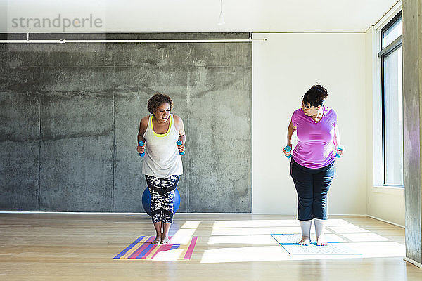Frau in voller Länge sieht Freundin an  die auf Übungsmatte steht  während sie Hanteln gegen die Wand im Yoga-Studio hält
