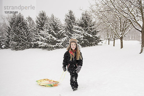 Mädchen in voller Länge in warmer Kleidung beim Spaziergang mit Schlitten auf verschneitem Feld gegen Bäume bei Schneefall