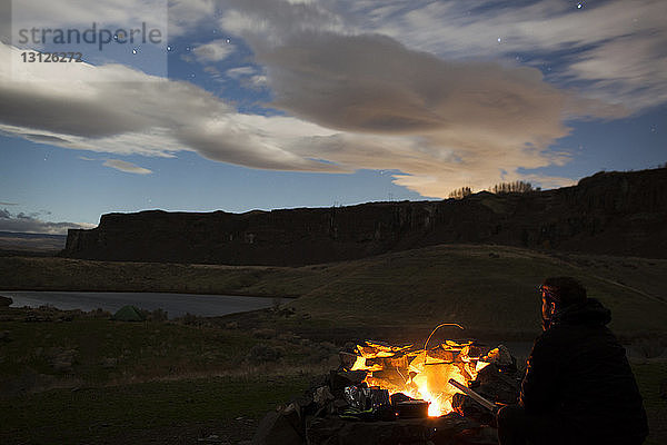Wanderer sitzt am Lagerfeuer vor bewölktem Himmel
