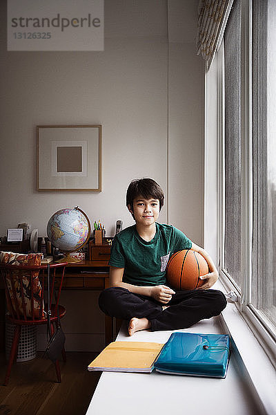 Porträt eines Jungen  der zu Hause am Fenster den Ball hält und auf dem Tisch sitzt
