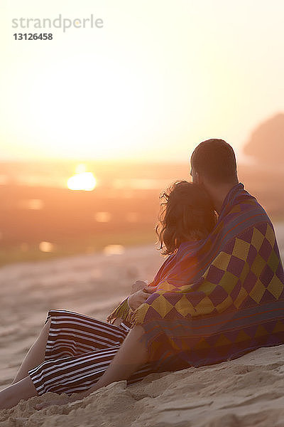 Nachdenkliches junges Paar in eine Decke gewickelt  während es bei Sonnenuntergang am Strand sitzt