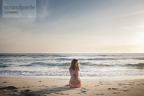 Rückansicht einer Frau  die bei Sonnenuntergang am Strand gegen den Himmel sitzt