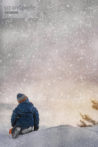 Rückansicht eines Jungen  der während des Schneefalls bei Sonnenuntergang auf einem schneebedeckten Feld sitzt