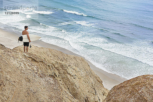 Mann trägt Surfbrett  während er auf einer Felsformation am Strand steht