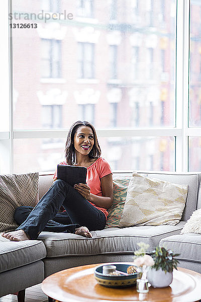 Glückliche Frau schaut weg  während sie zu Hause einen Tablet-Computer auf dem Sofa gegen das Fenster hält