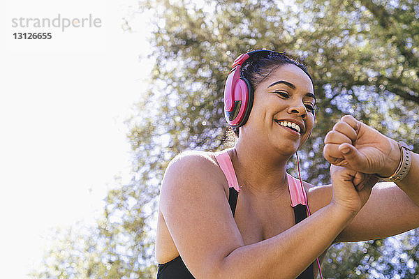 Niedriger Blickwinkel einer glücklichen Frau  die beim Musikhören im Park auf den Fitnesstracker schaut