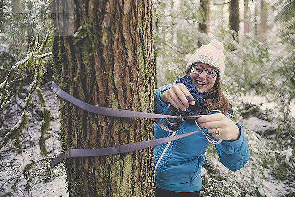 Lächelnde Frau bindet im Wald im Lynn Canyon Park im Winter Hängemattenbänder an Baumstamm