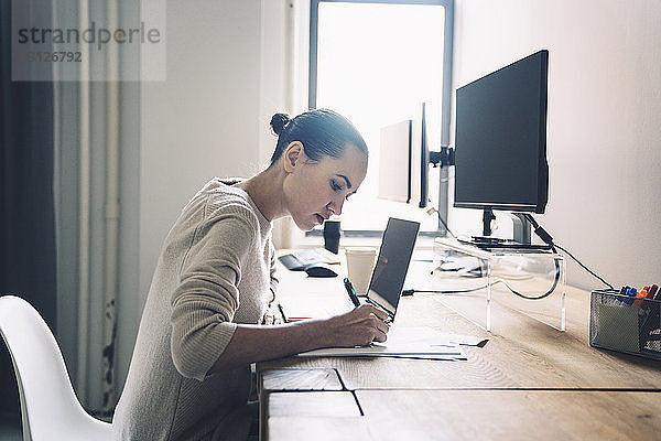 Seitenansicht einer Geschäftsfrau  die schreibt  während sie im Büro am Schreibtisch sitzt