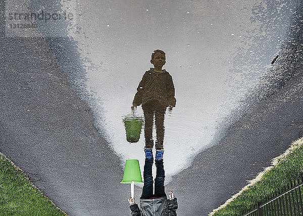 Auf dem Kopf stehendes Bild eines Jungen  der auf nasser Straße steht und einen Eimer trägt