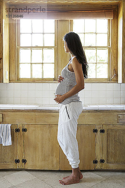 Nachdenkliche schwangere Frau berührt Bauch  während sie in der Küche steht