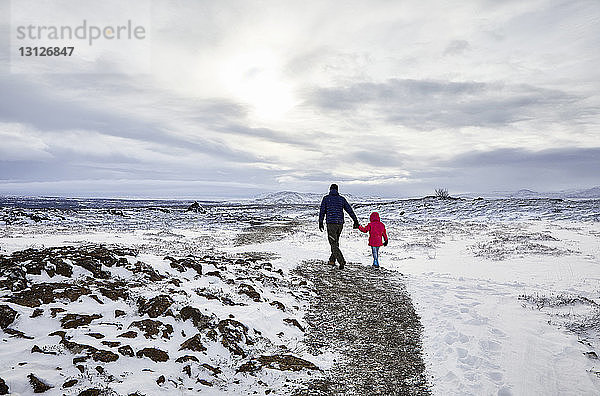 Rückansicht eines Vaters und einer Tochter  die sich an den Händen halten  während sie über eine schneebedeckte Landschaft vor bewölktem Himmel gehen