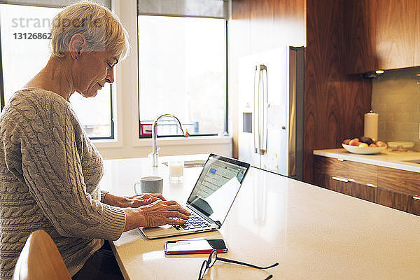 Seitenansicht einer älteren Frau  die am Laptop tippt  während sie am Küchentisch sitzt