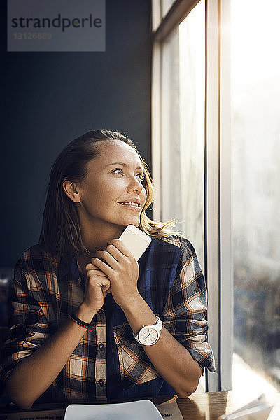 Lächelnde Frau hält Smartphone in der Hand  während sie durchs Fenster in ein Café schaut