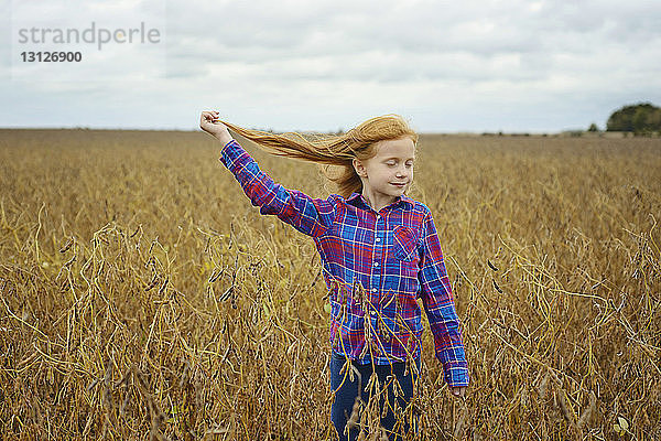 Unbekümmertes Mädchen mit geschlossenen Augen hält ihr Haar  während sie inmitten eines Sojabohnenfeldes gegen den Himmel steht
