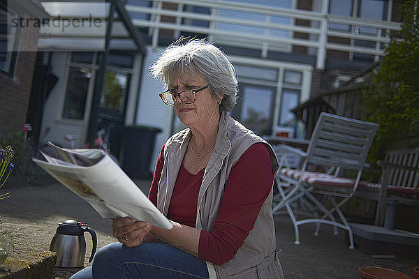 Ältere Frau liest Zeitschrift  während sie auf dem Feld im Hof sitzt