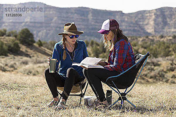 Freundinnen schauen sich ein Buch an  während sie bei Sonnenschein auf Campingstühlen auf einem Feld gegen die Berge sitzen