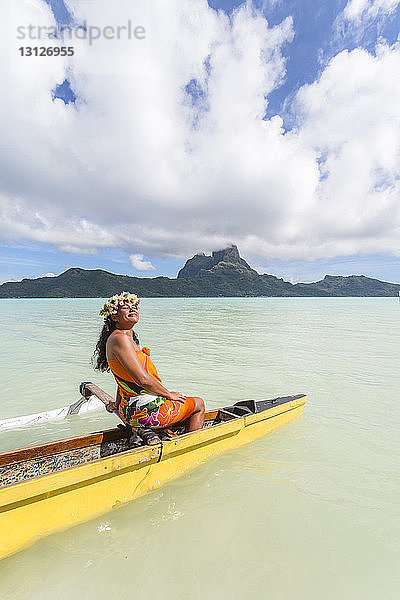 Frau in traditioneller Kleidung beim Kanufahren auf der Lagune der Insel Bora Bora vor bewölktem Himmel